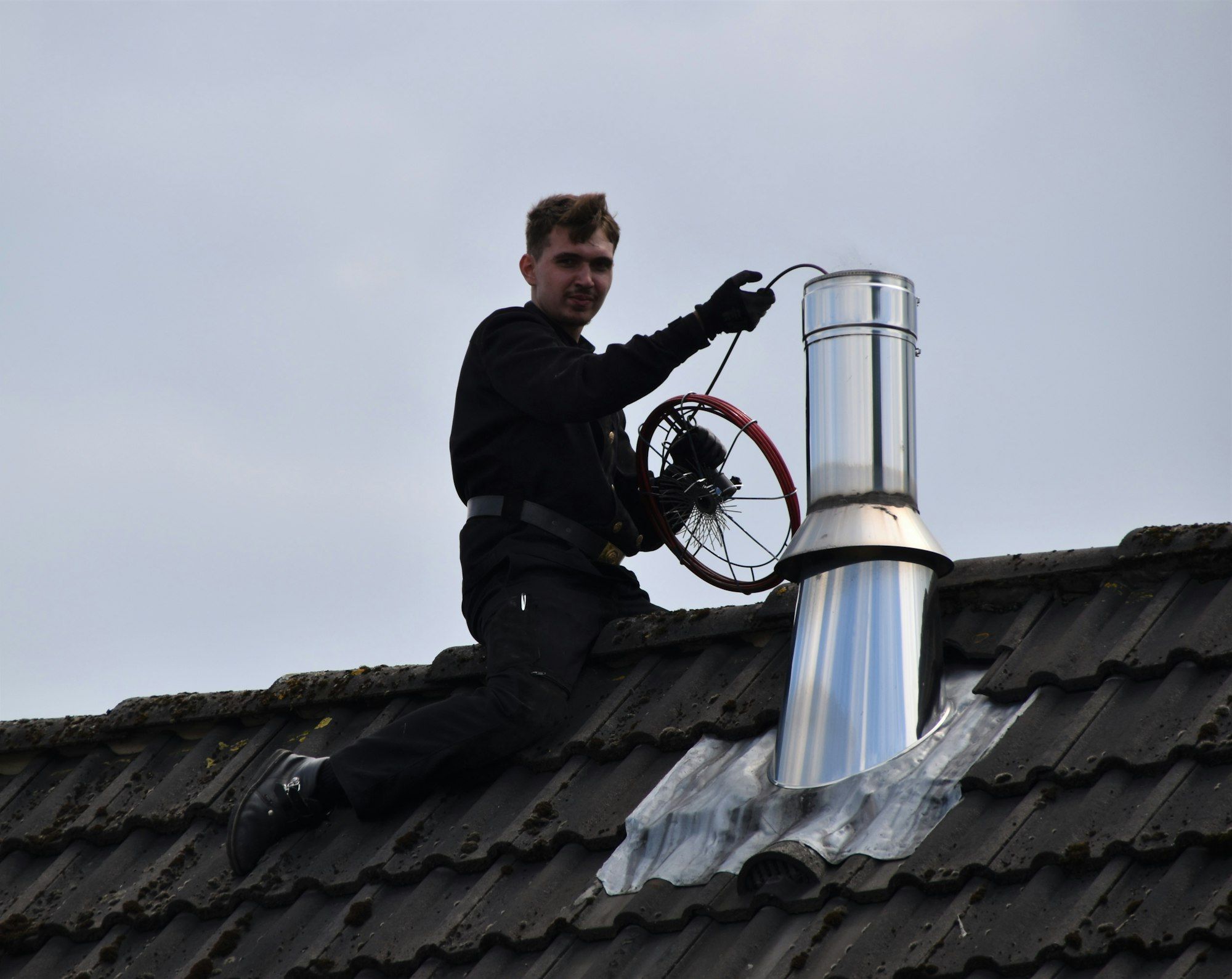 Chimney sweep working at a steel chimney pipe on a dark shingle roof
