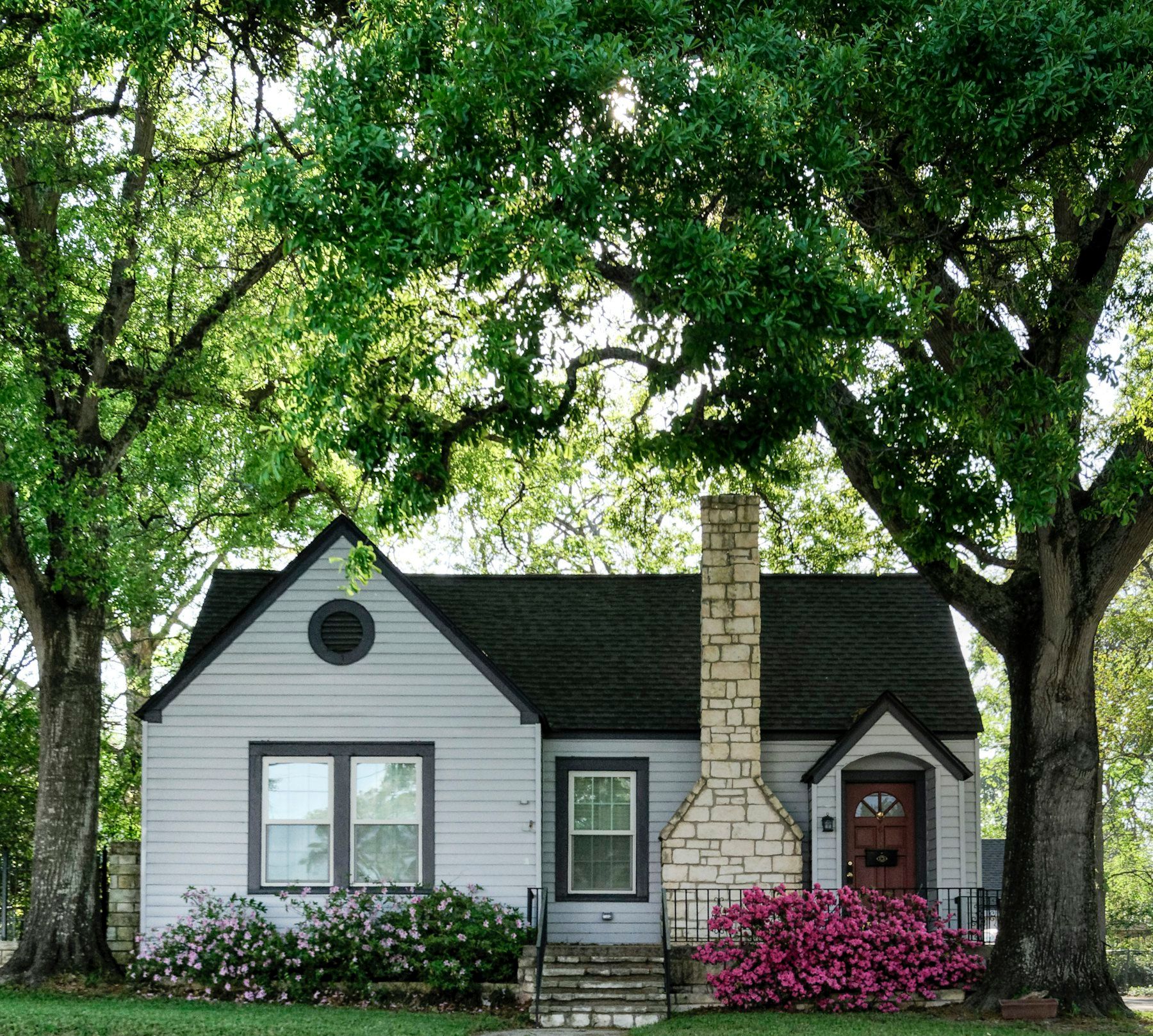 Small white house with a stone chimney and pink flowers under tall trees